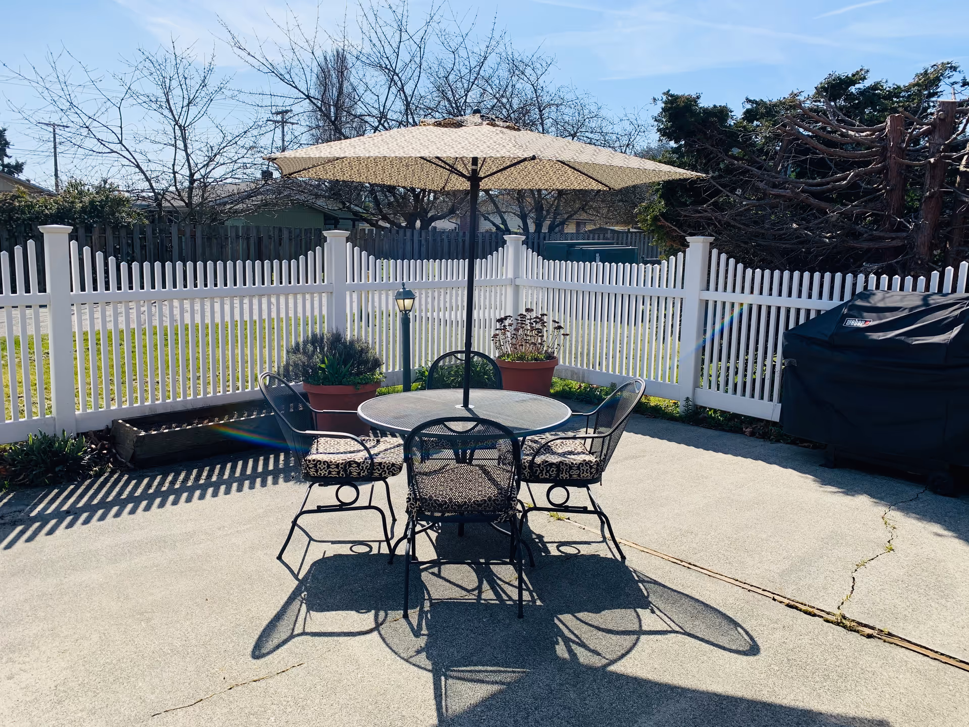 Outdoor patio with round table, umbrella, and metal chairs enclosed by a white fence.