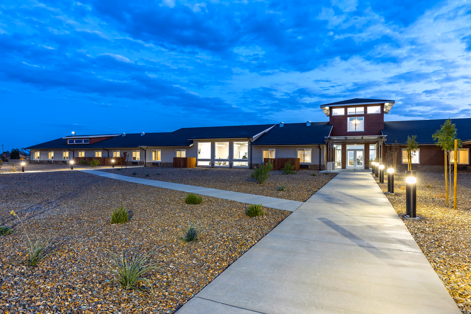 Exterior view of The Landings Of Prescott Valley facility at dusk with a well-lit entrance, a concrete walkway lined with lights, and a landscaped area with small plants and gravel.