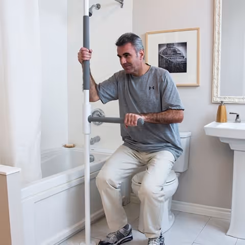 A man in a gray shirt uses a bathroom safety grab bar beside a bathtub.