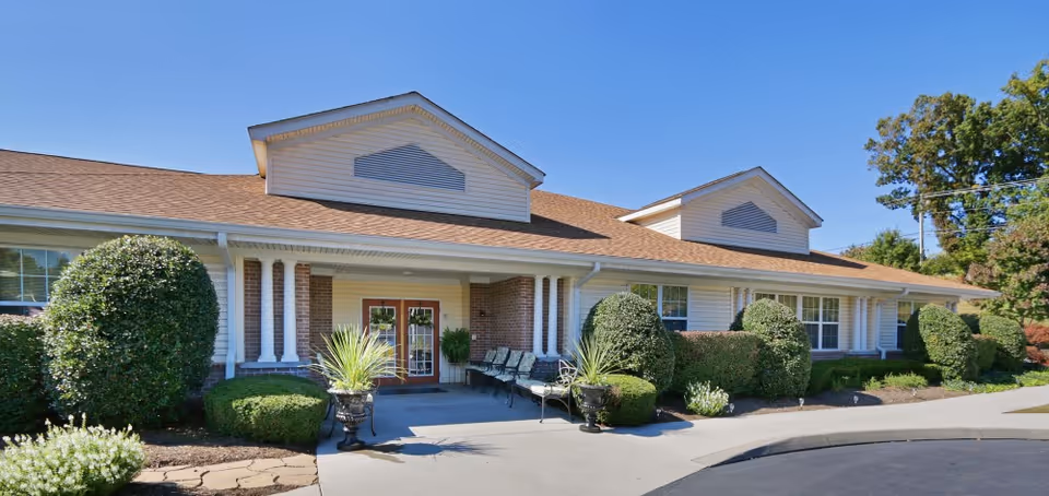 Exterior front view of a single-story building with a brown shingled roof, white siding, and brick accents. The entrance features double glass doors with white columns on either side, surrounded by neatly trimmed bushes and potted plants. There is a paved driveway and a clear blue sky above.