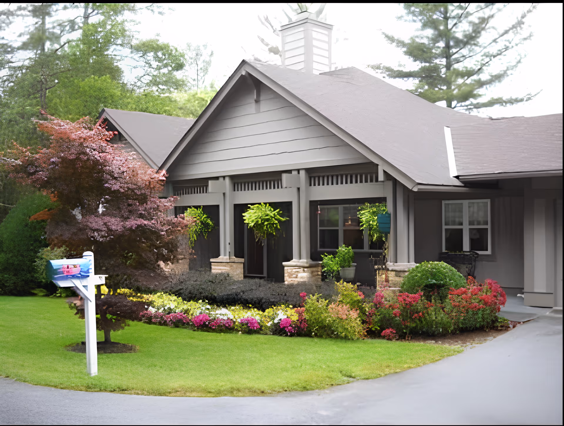 Exterior view of a single-story building with a gray roof and siding, surrounded by a well-maintained garden with colorful flowers and green shrubs. A white mailbox with a blue and pink design is visible on the left side of the image, and there are trees in the background.