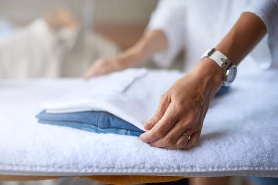 Hands fold a white towel on a bed in a softly lit room.