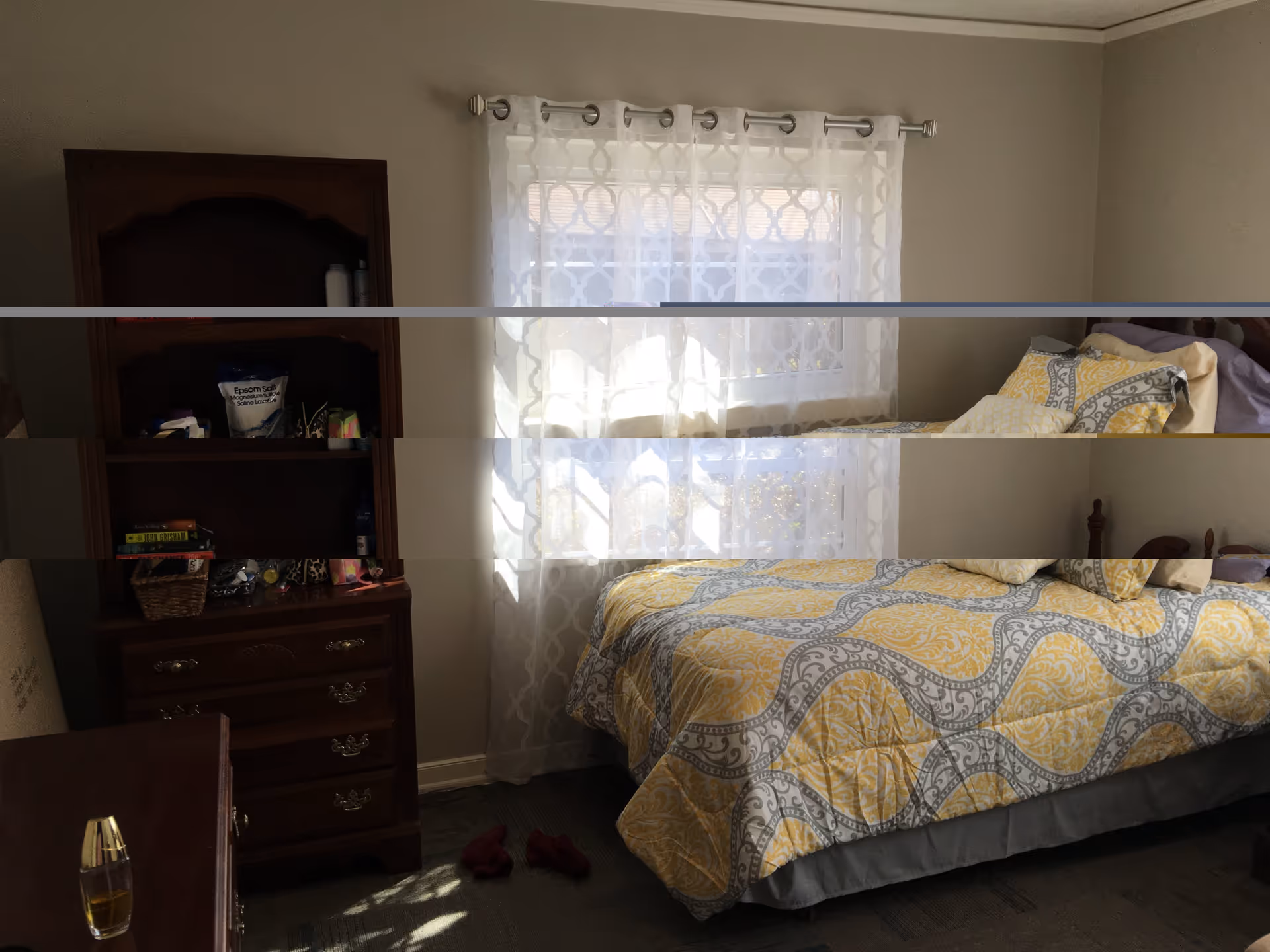A cozy bedroom with a bed covered in a yellow and gray patterned comforter and multiple pillows. There is a wooden dresser with shelves on the left side holding various items, and a window with sheer white curtains letting in natural light behind the bed.