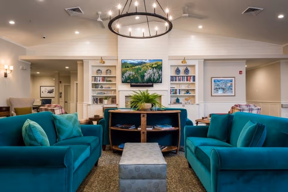 Communal lounge with teal sofas arranged around a central ottoman and console, a TV above a fireplace, and built-in bookshelves.