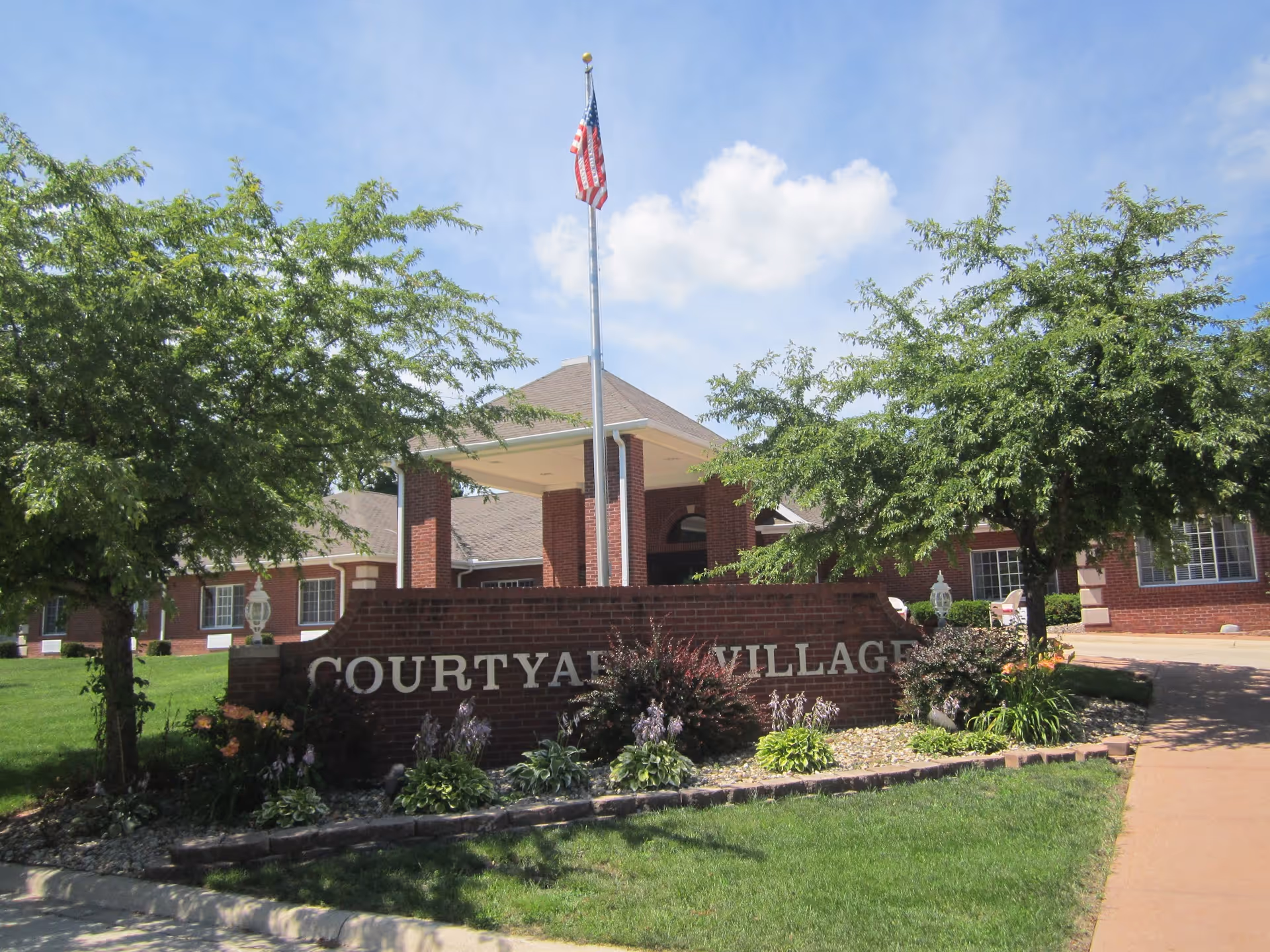 Front entrance of a red-brick senior living building with a brick sign reading COURTYARD VILLAGE, flagpole, canopy, and landscaped trees.