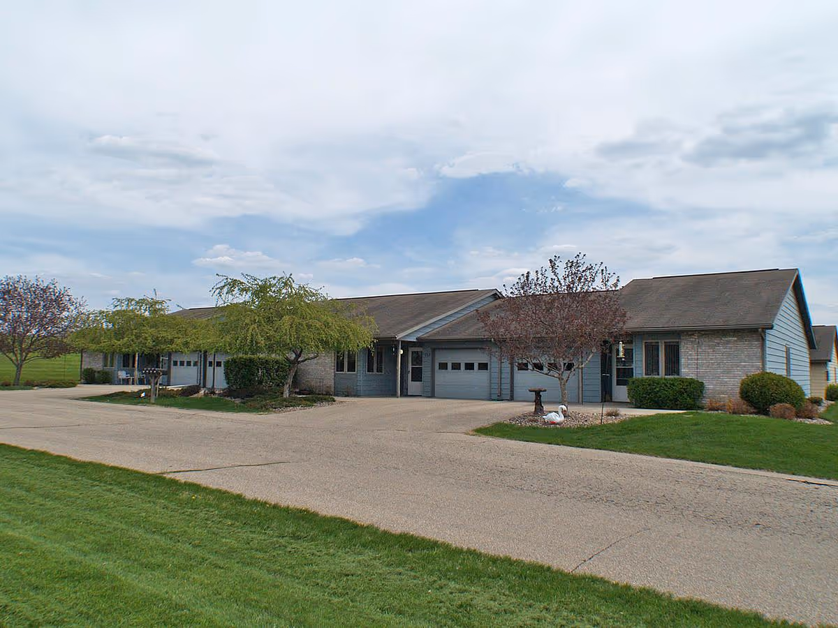 Single-story townhome units with attached garages, driveways, and landscaped front lawns under a cloudy sky.