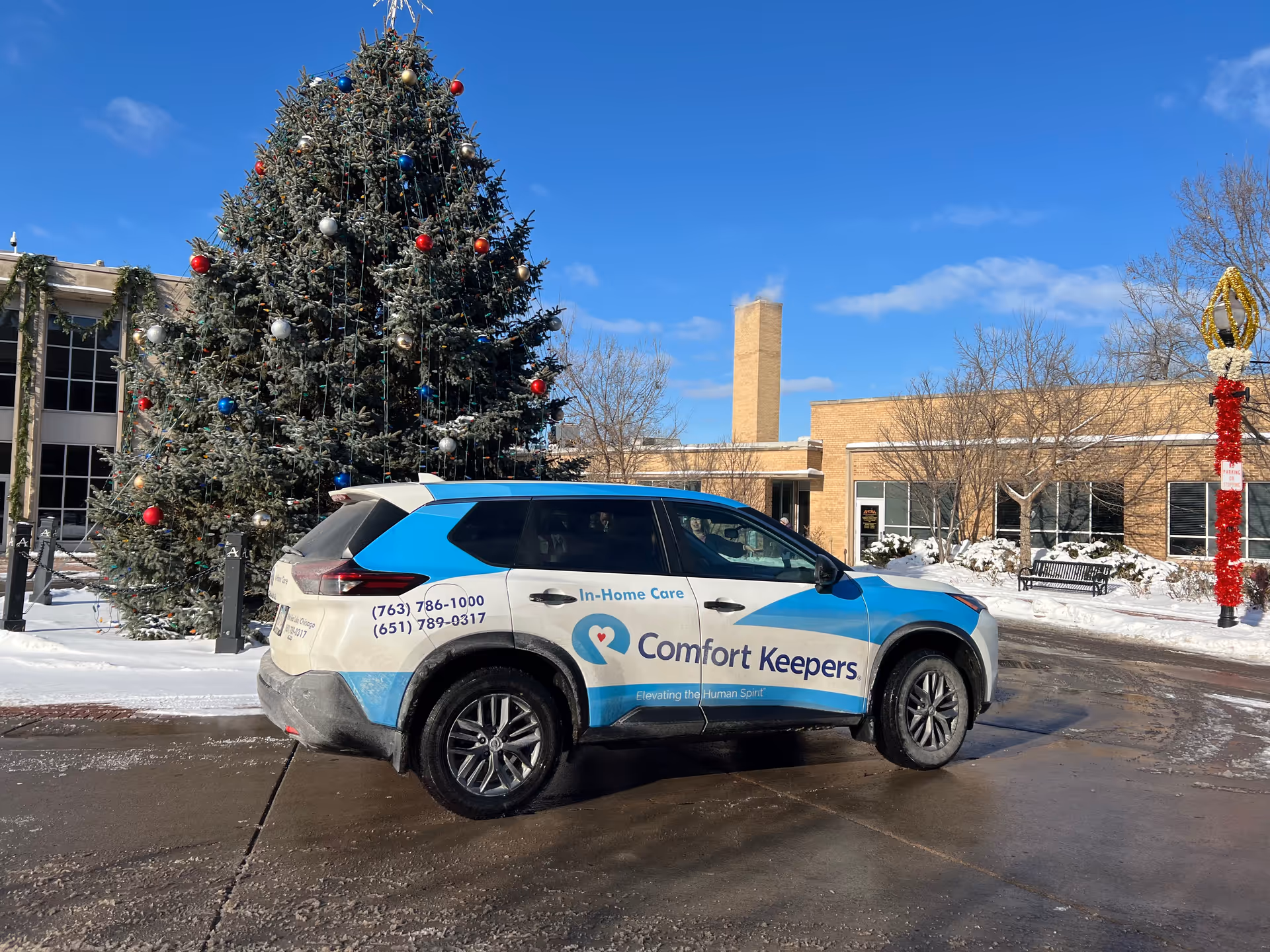 A Comfort Keepers vehicle parked outside a brick building beside a large Christmas tree.