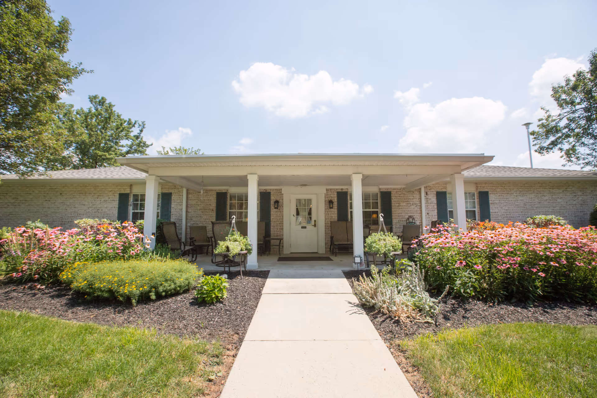 Front exterior view of a single-story brick building with a covered porch, several chairs, hanging plants, and well-maintained flower beds on either side of a concrete walkway leading to a white door under a clear blue sky.