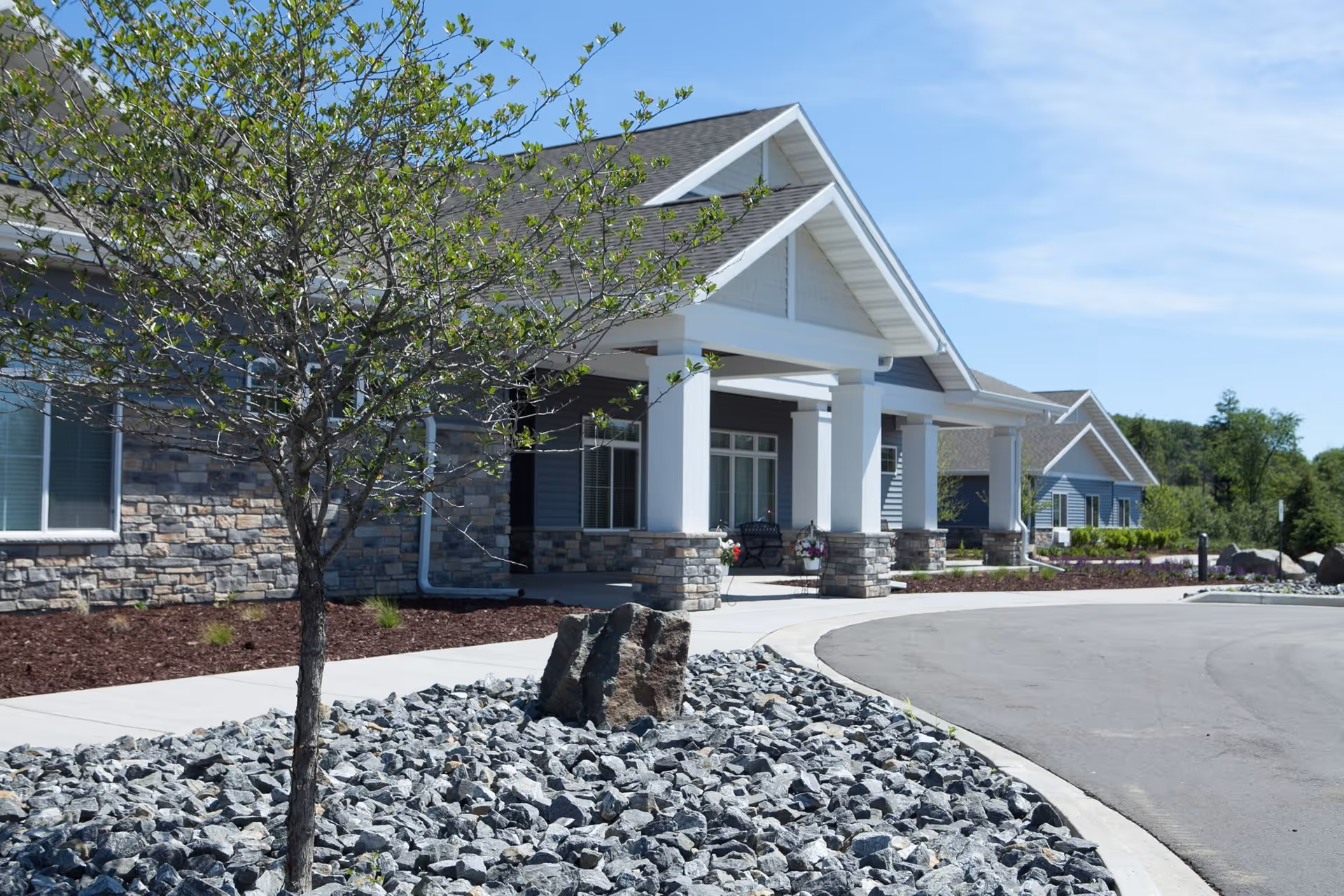 Exterior view of Weston Memory Care facility showing a covered entrance with white pillars and stone accents, a small tree planted in a rock bed, and a curved driveway under a clear blue sky.