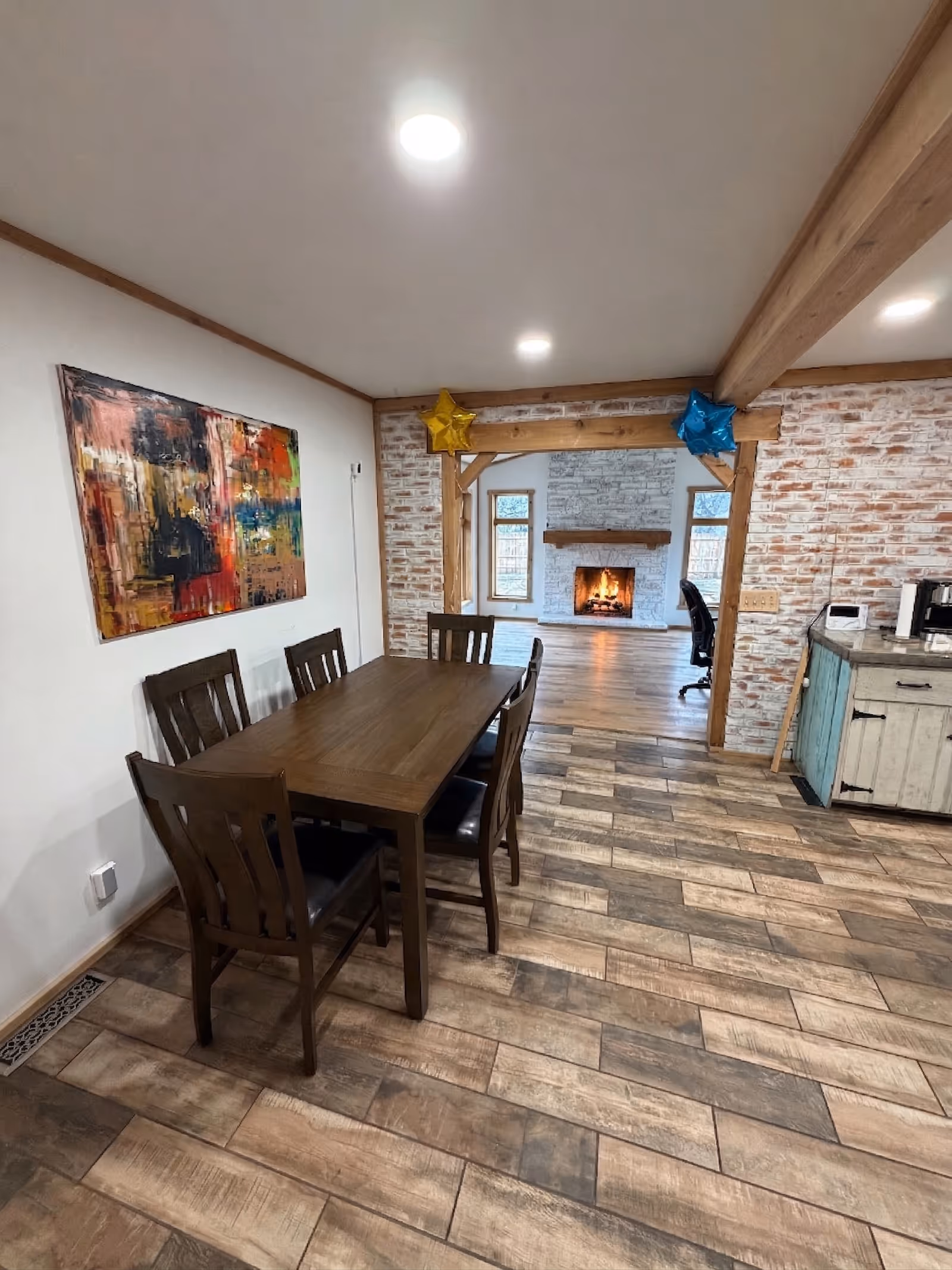 Wooden dining table with six chairs in a brick-walled dining area, fireplace in background.