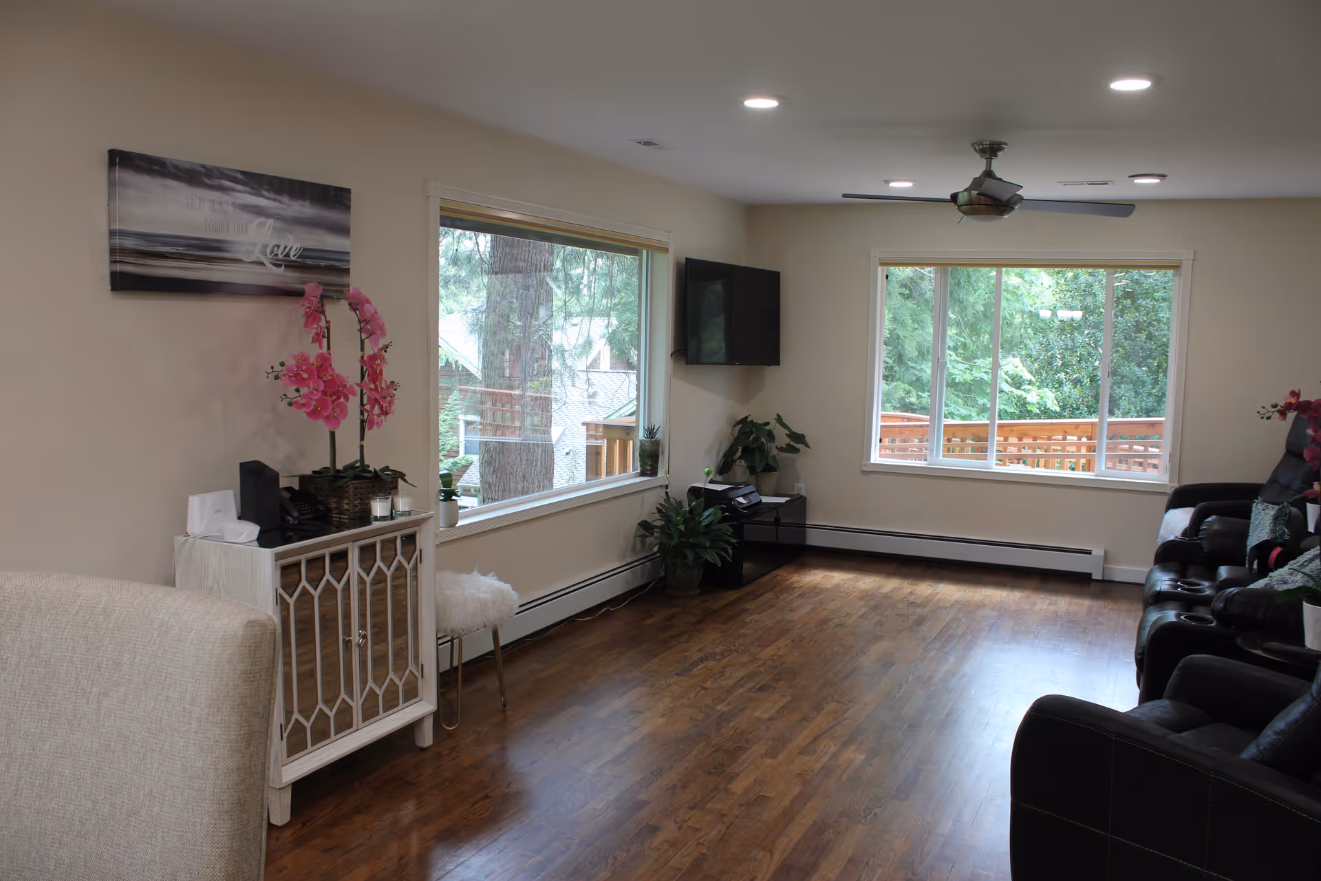 A bright senior living lounge with sofas, windows, a wall-mounted TV, and wood floor.