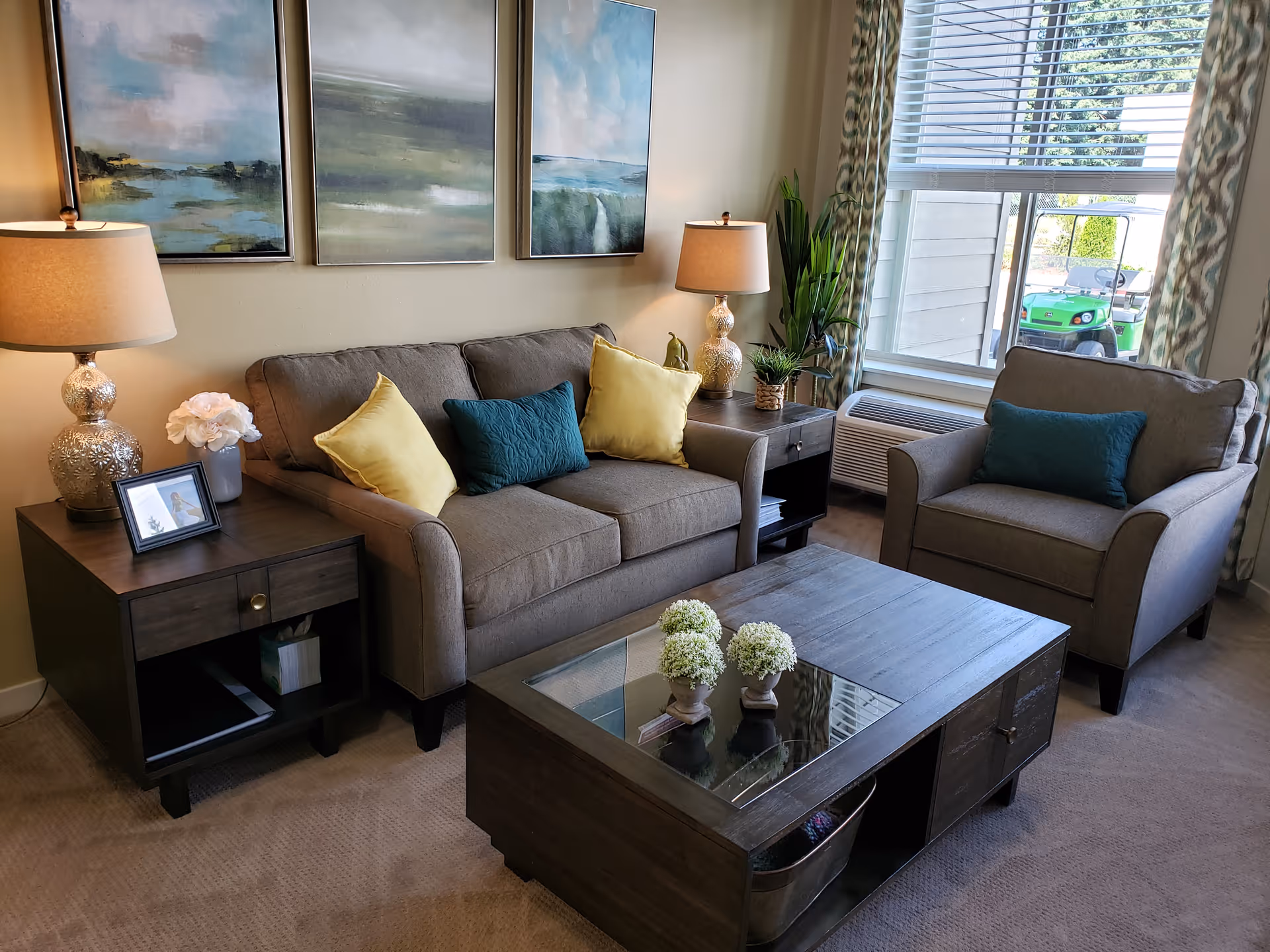 A cozy living room with a gray sofa and matching armchair, both adorned with colorful throw pillows. There are two side tables with decorative lamps and plants, a coffee table with small flower arrangements, and three landscape paintings on the wall. A large window with patterned curtains lets in natural light.