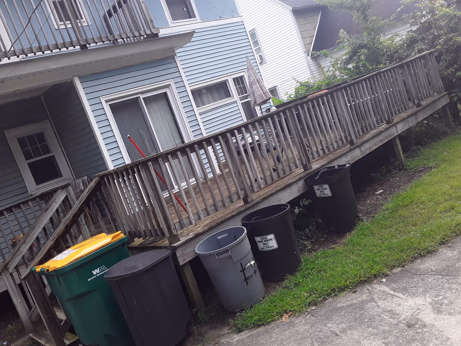 Backyard view of a blue house with a wooden deck and several trash cans.