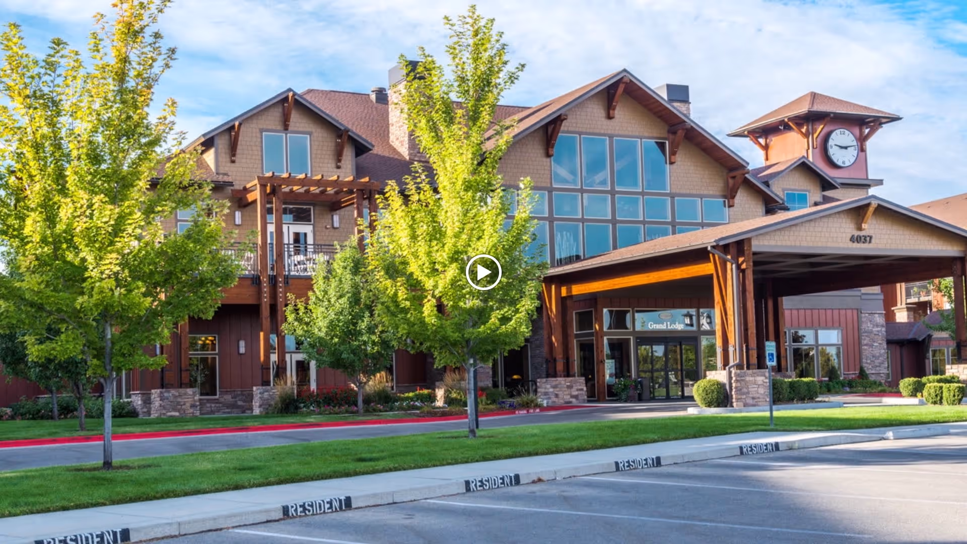 Exterior view of a senior living facility named Touchmark at Meadow Lake Village, featuring a large building with a clock tower, multiple windows, and a covered entrance labeled 'Grand Lodge'. The foreground shows a parking area with 'Resident' parking spaces and green trees and grass around the building.