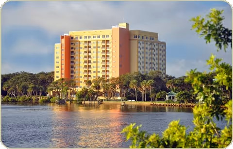 A tall multi-story building with balconies, situated near a body of water with trees and greenery surrounding the area under a partly cloudy sky.