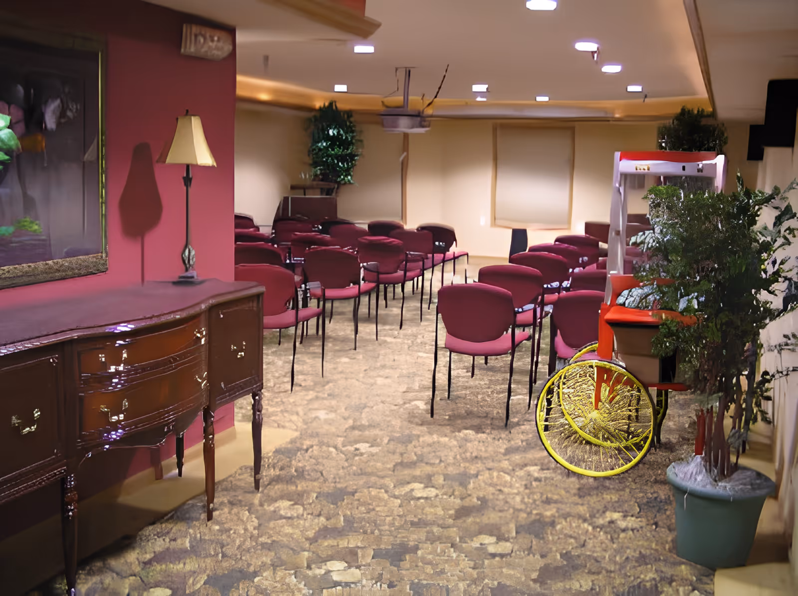Interior room with rows of maroon chairs arranged facing forward, a wooden sideboard with a lamp on the left, potted plants on the right, and a popcorn machine near the back wall.