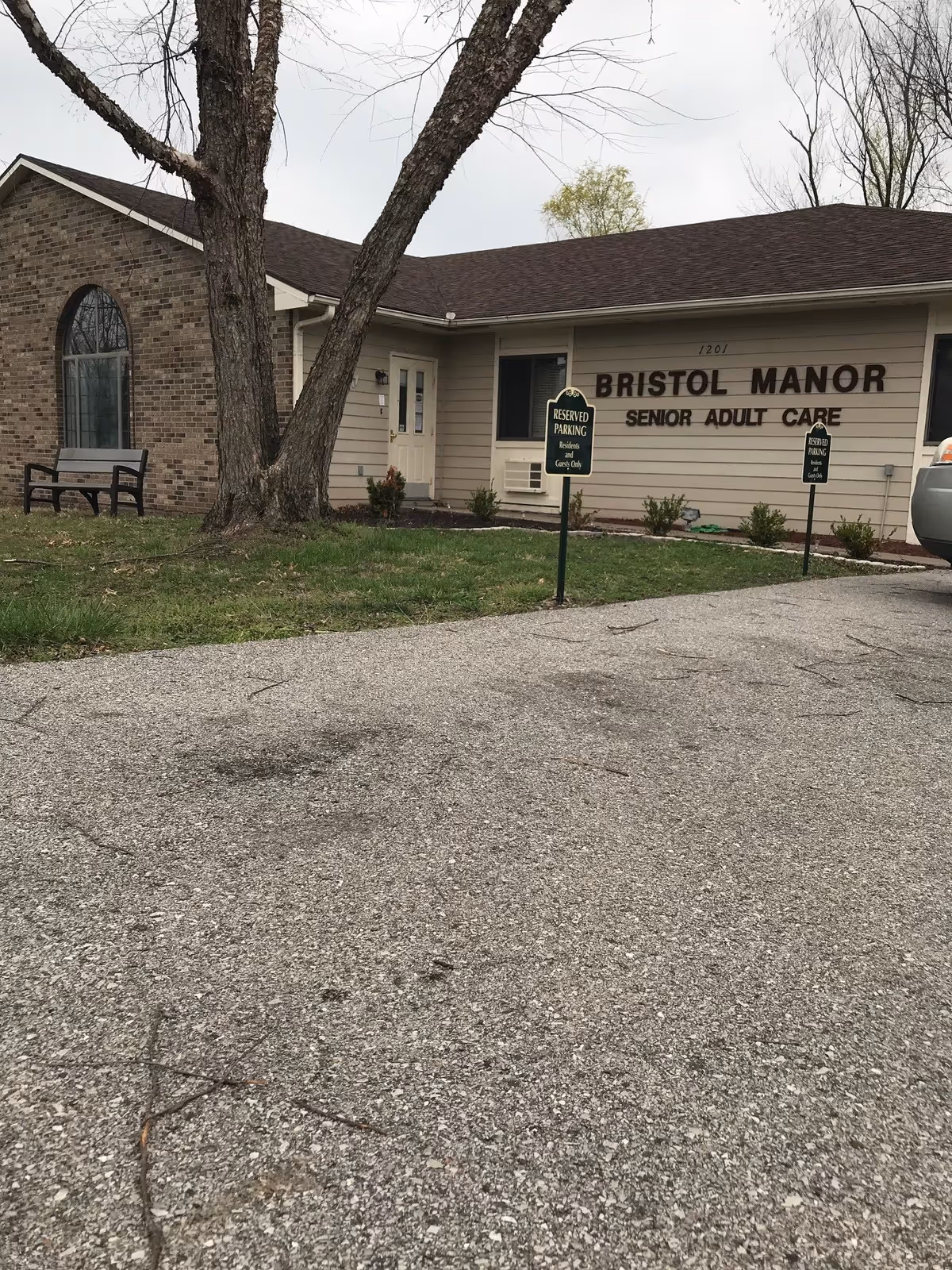 Front exterior of Bristol Manor senior adult care building with parking signs, a bench, and a large tree.