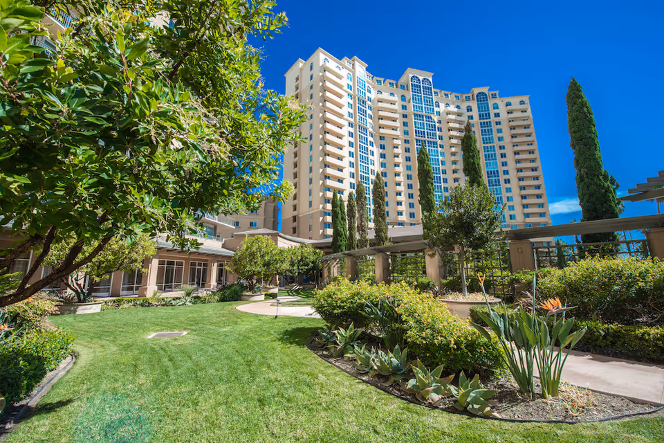 Tall beige senior living building with landscaped courtyard and bright blue sky.