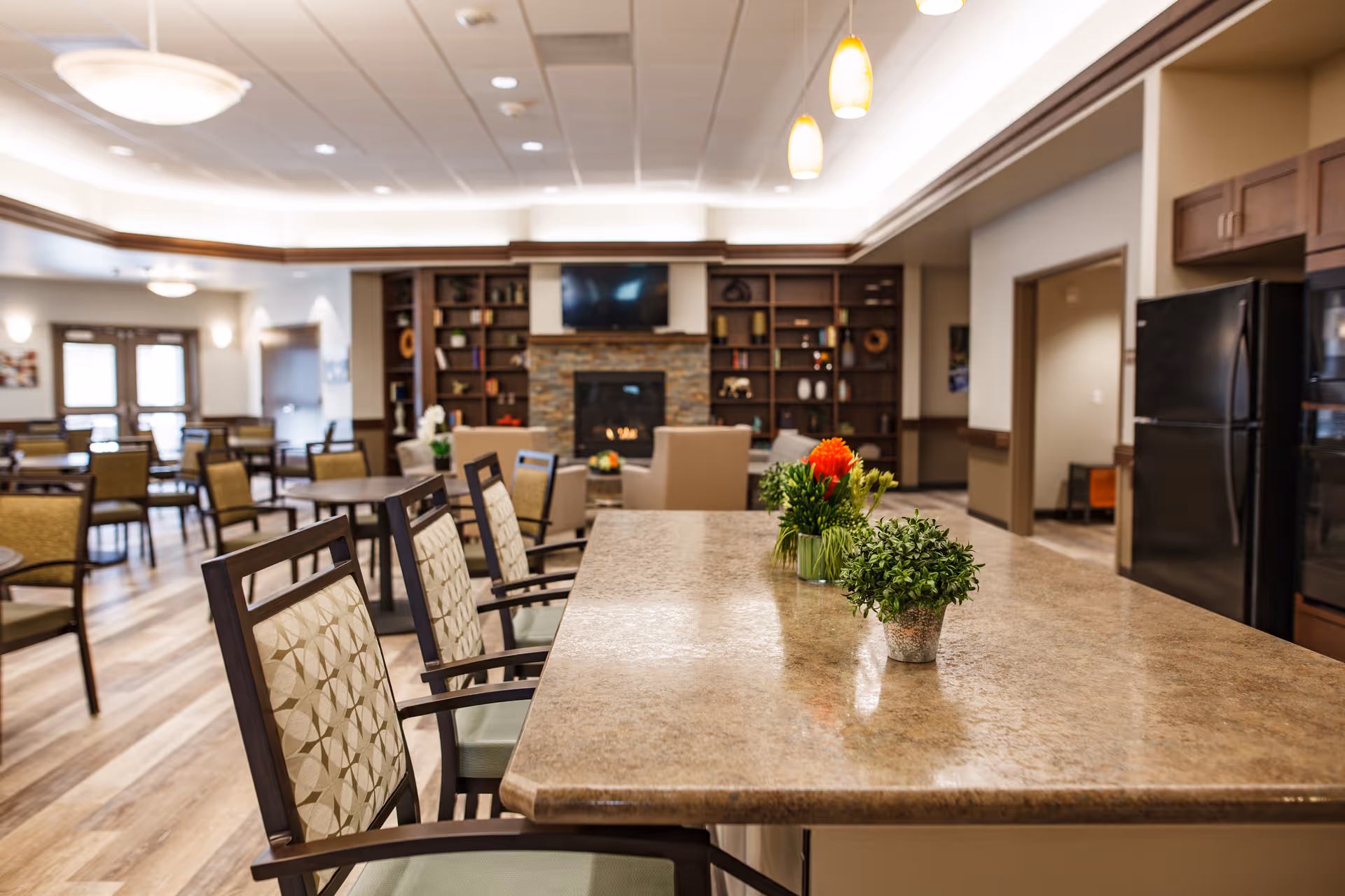 Interior view of a senior living facility common area with a large countertop island adorned with small potted plants, several chairs around the island, wooden flooring, a fireplace with a TV mounted above it, built-in bookshelves, and multiple tables and chairs in the background.