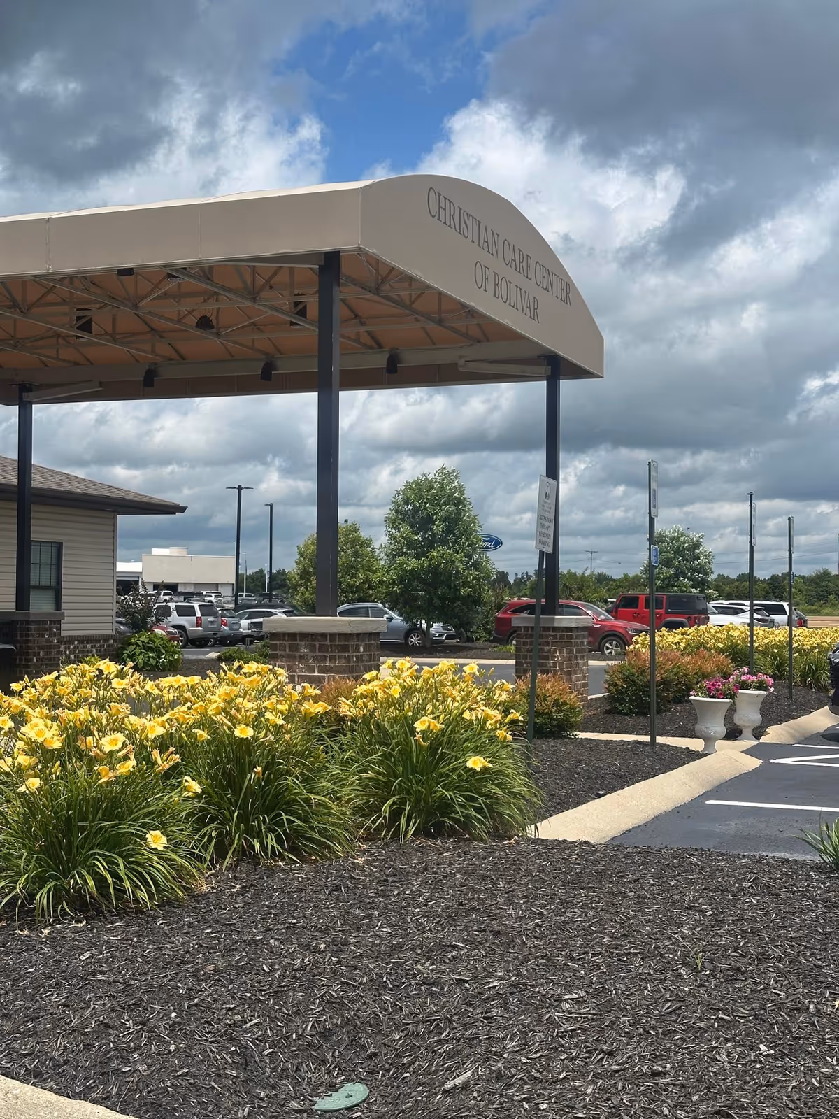 Exterior entrance canopy of Christian Care Center of Bolivar with parked cars and yellow flowers.
