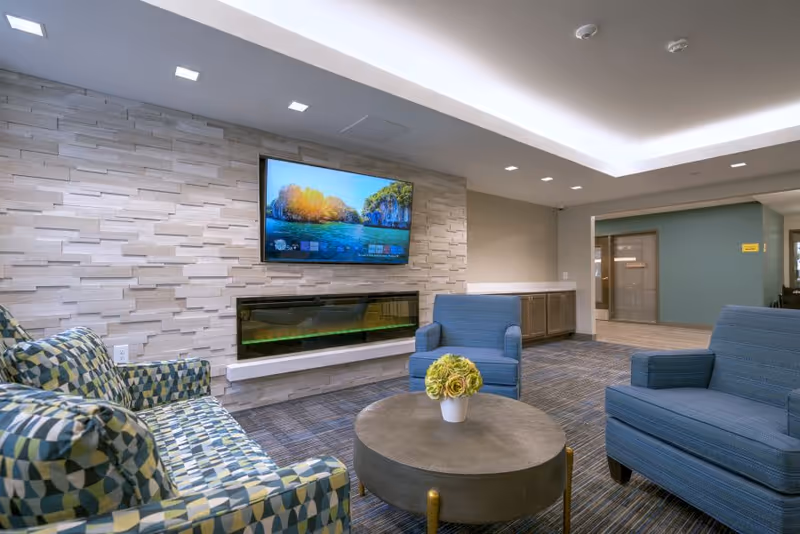 Modern lounge with patterned sofa, blue armchairs, a round coffee table with flowers, wall-mounted TV and electric fireplace.