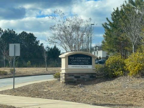 Stone entrance sign reading "The Landings of Canton Hills" by a roadside with trees and cloudy sky.