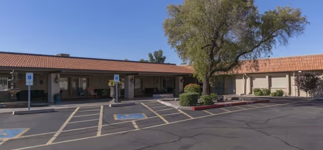 Exterior view of Desert Blossom Health and Rehabilitation facility showing a single-story building with a red-tiled roof, several handicap parking spaces in front, a large tree, and some shrubs and benches near the entrance under a clear blue sky.