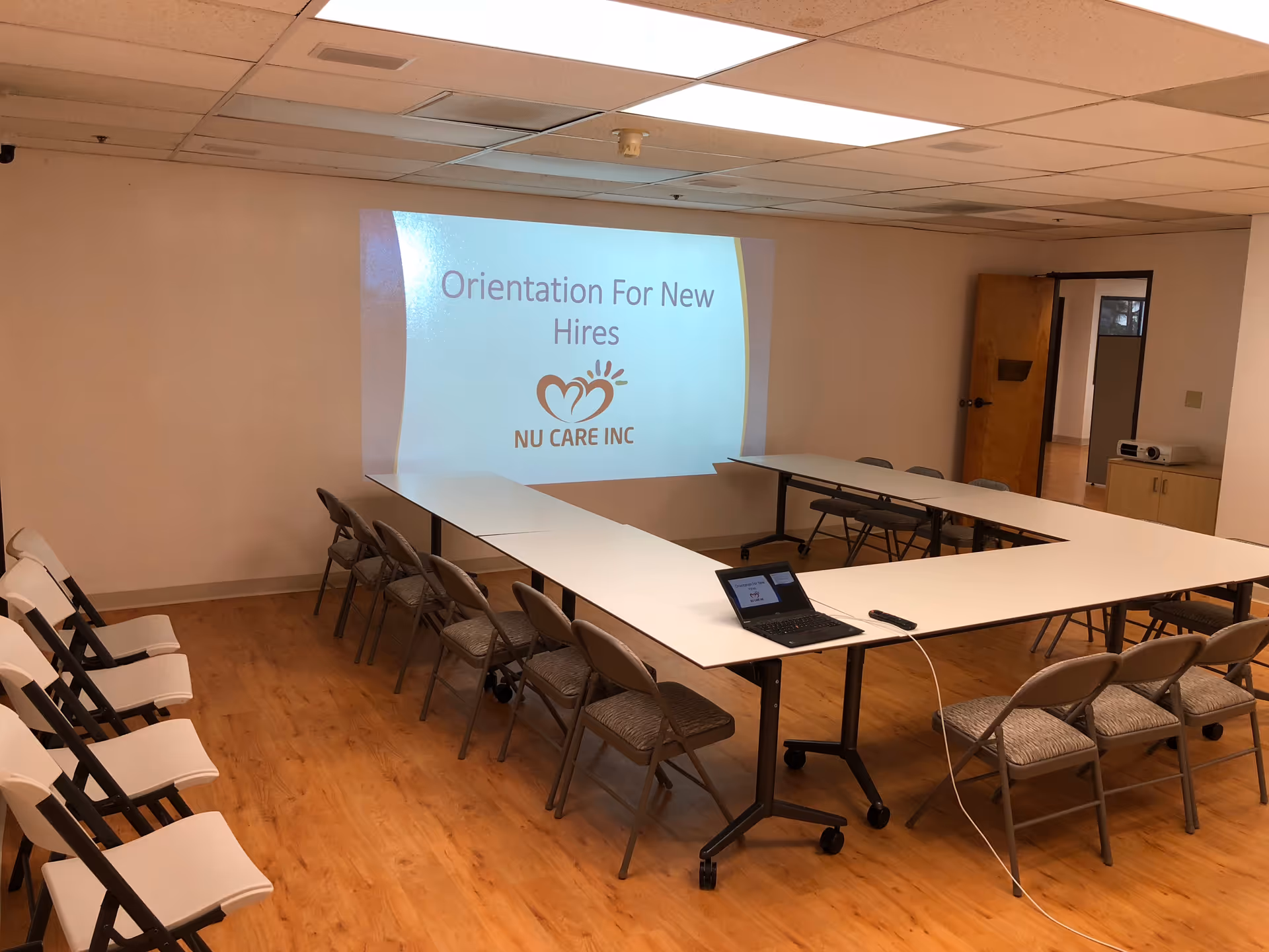 Empty conference room with U-shaped tables, chairs, and a projector screen displaying Nu Care logo.