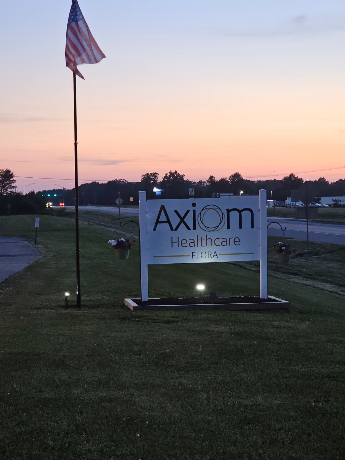 Outdoor view of a sign for Axiom Healthcare Flora on a grassy area near a road at dusk, with an American flag on a flagpole beside it and a sunset sky in the background.