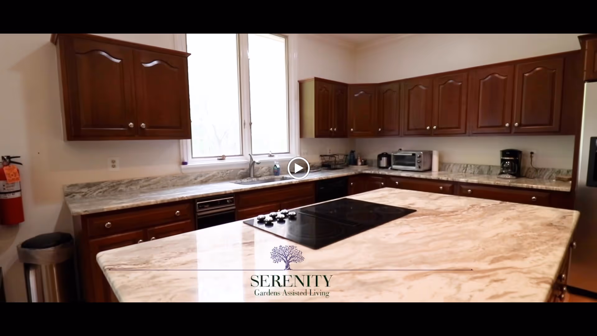 A spacious kitchen with marble countertops and dark wooden cabinets. The kitchen features a stovetop on a large island, a toaster oven, a coffee maker, a dishwasher, and a stainless steel refrigerator. There is a window above the sink letting in natural light. A fire extinguisher and trash can are visible on the left side.