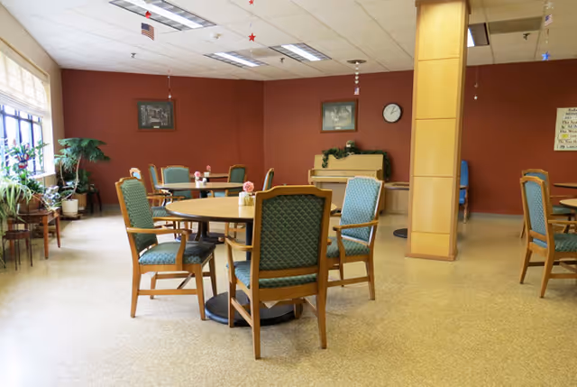 Communal dining/activity room with round tables and green upholstered chairs, plants, a piano and a wall clock.