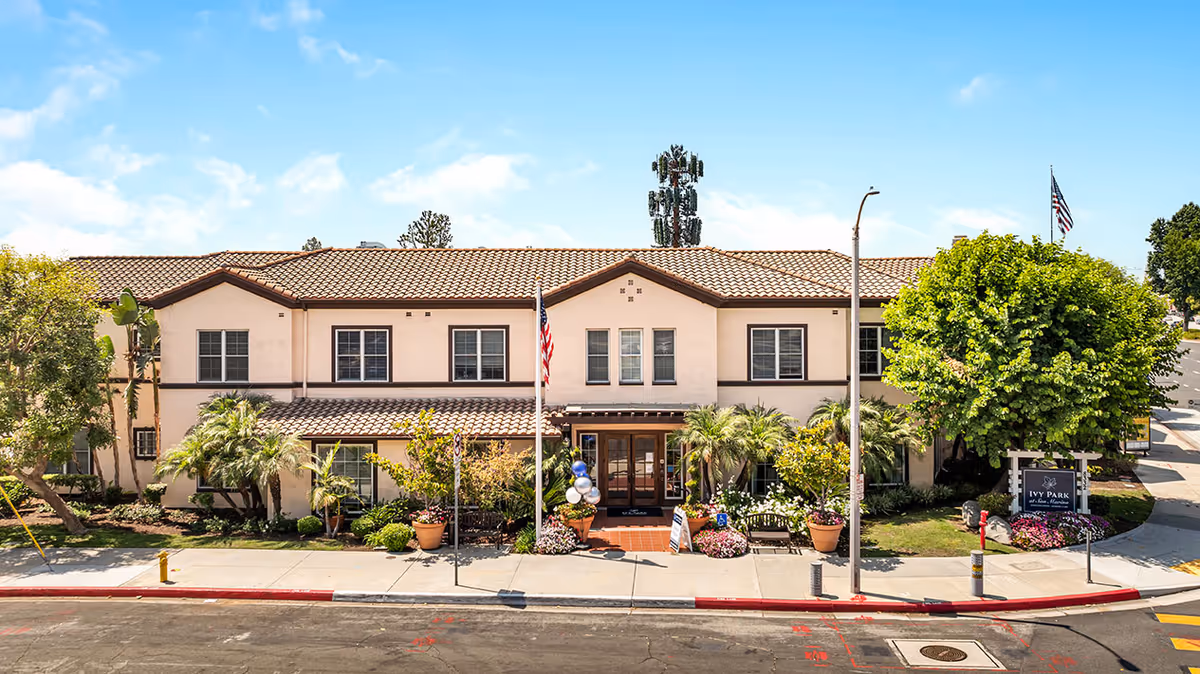 Front exterior view of a two-story senior living facility building with a tiled roof, multiple windows, and a central entrance. The entrance is flanked by potted plants and benches, with an American flag on a pole in front. There are trees and landscaping around the building, and a sign on the right side reads 'Ivy Park at San Marino'. The sky is clear and blue.