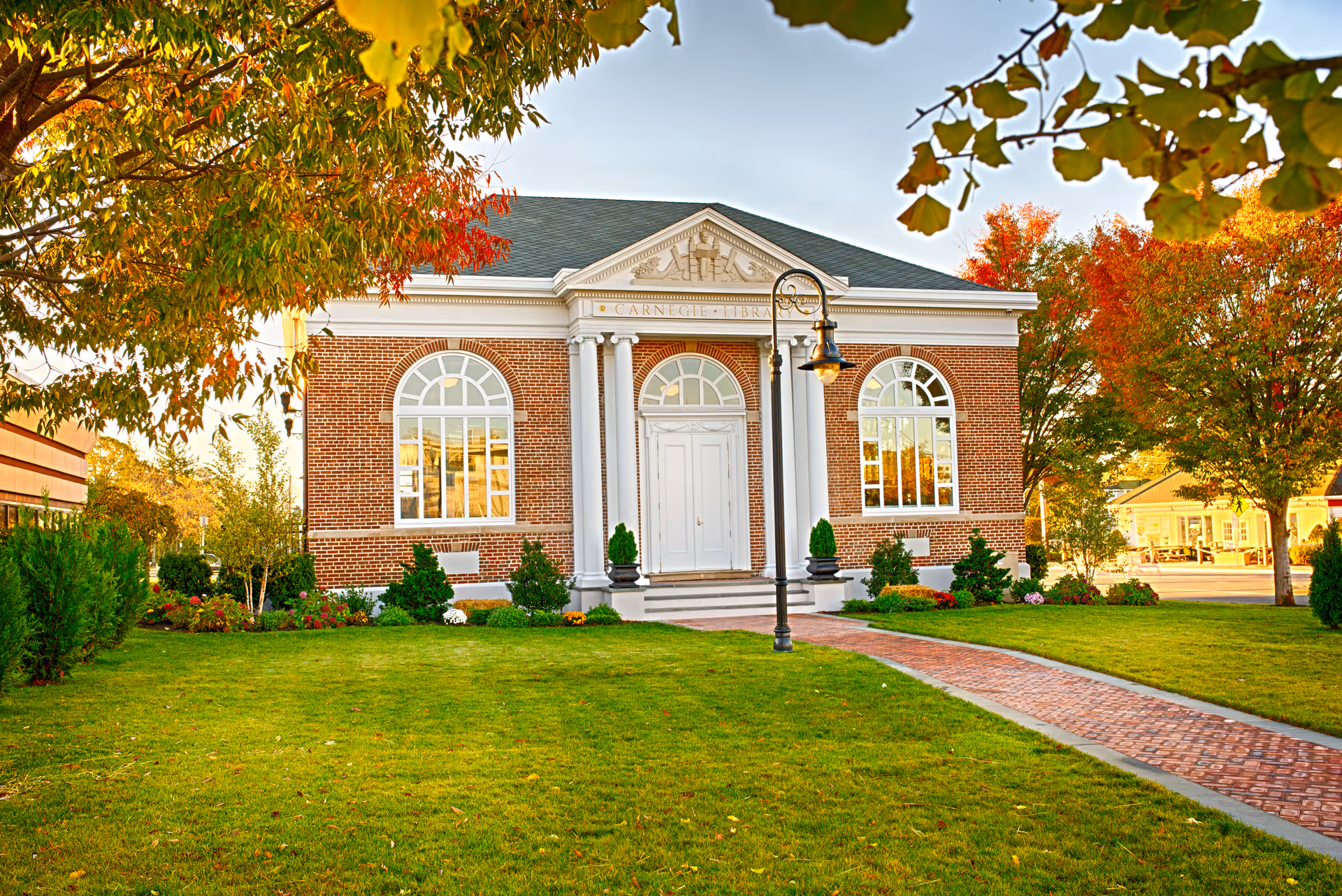 Brick colonial-style community building with white columns and arched windows, a brick walkway, and autumn trees on a manicured lawn.
