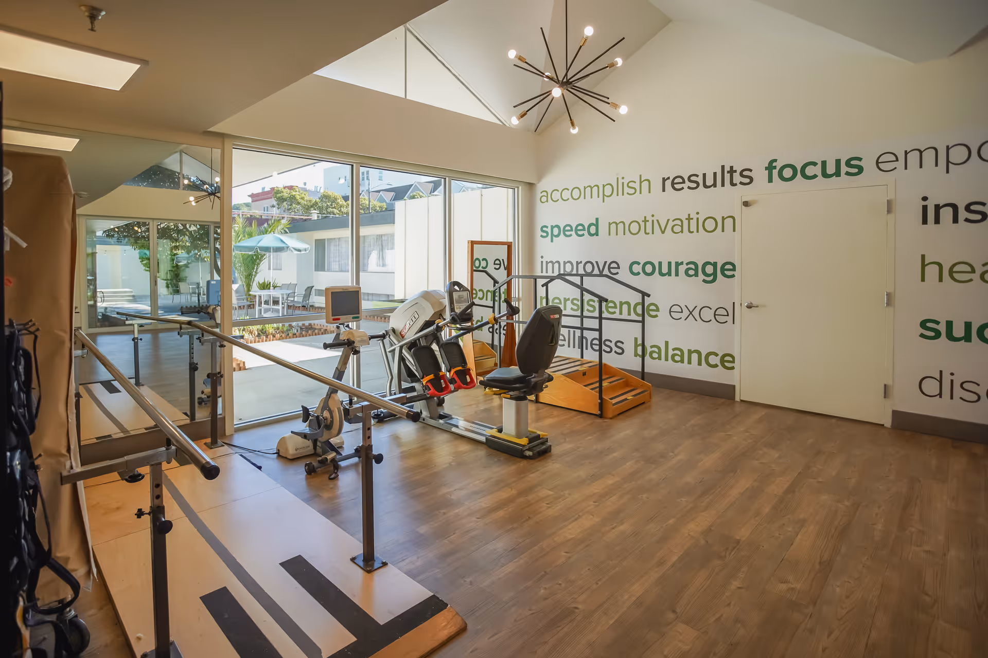 A bright rehabilitation room with wooden flooring, parallel bars for walking exercises, a recumbent exercise bike, and a small set of stairs for physical therapy. Large windows provide a view of an outdoor patio area with tables and umbrellas. The walls have motivational words like accomplish, results, focus, speed, motivation, improve, courage, persistence, wellness, and balance.