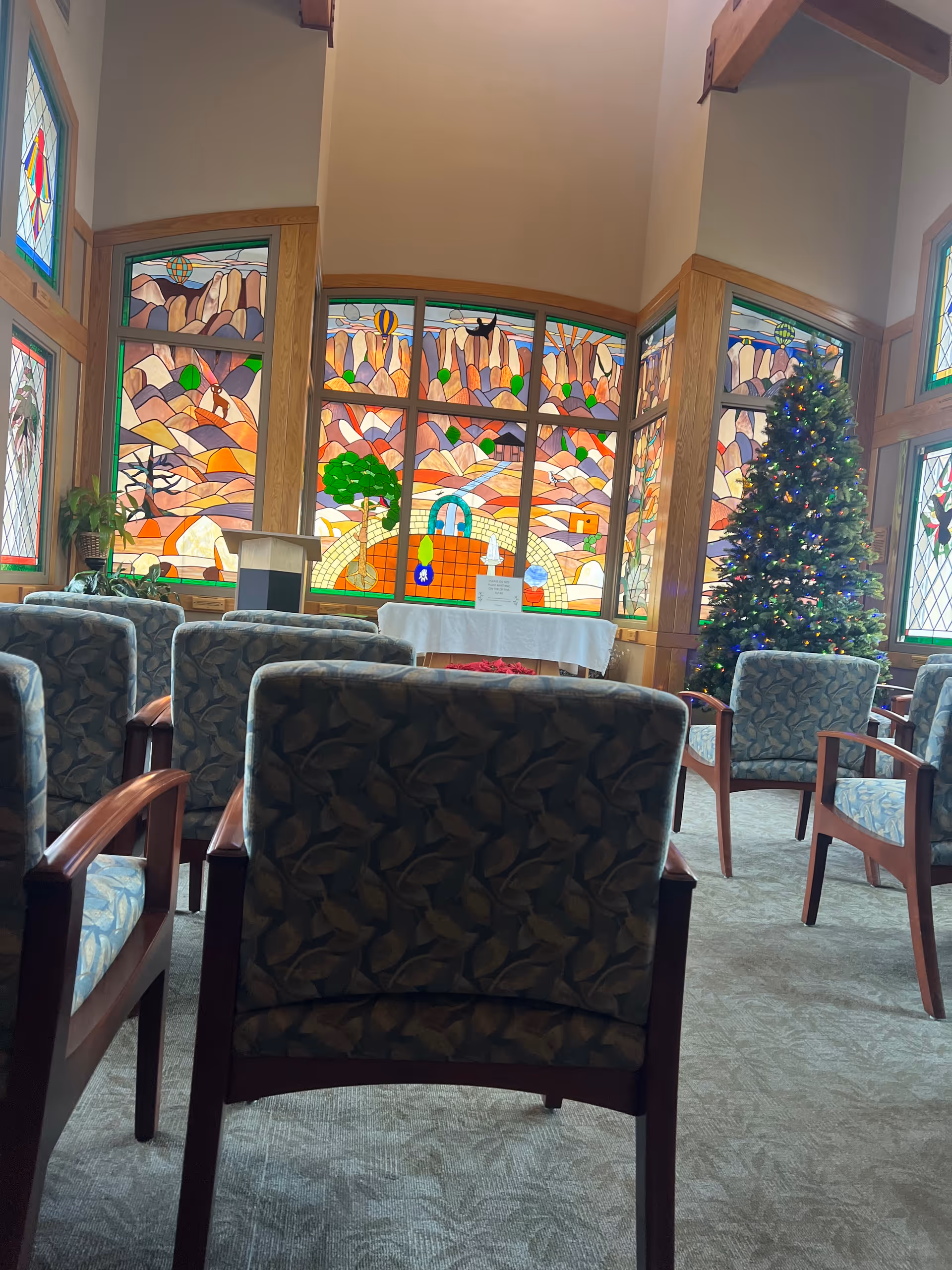 Interior lounge with patterned chairs, large stained glass windows, and a Christmas tree.