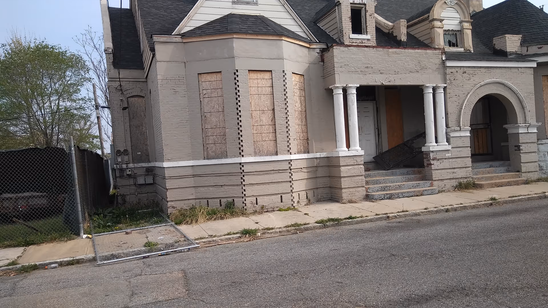 Old abandoned building exterior with boarded windows, broken porch, and fallen fence.