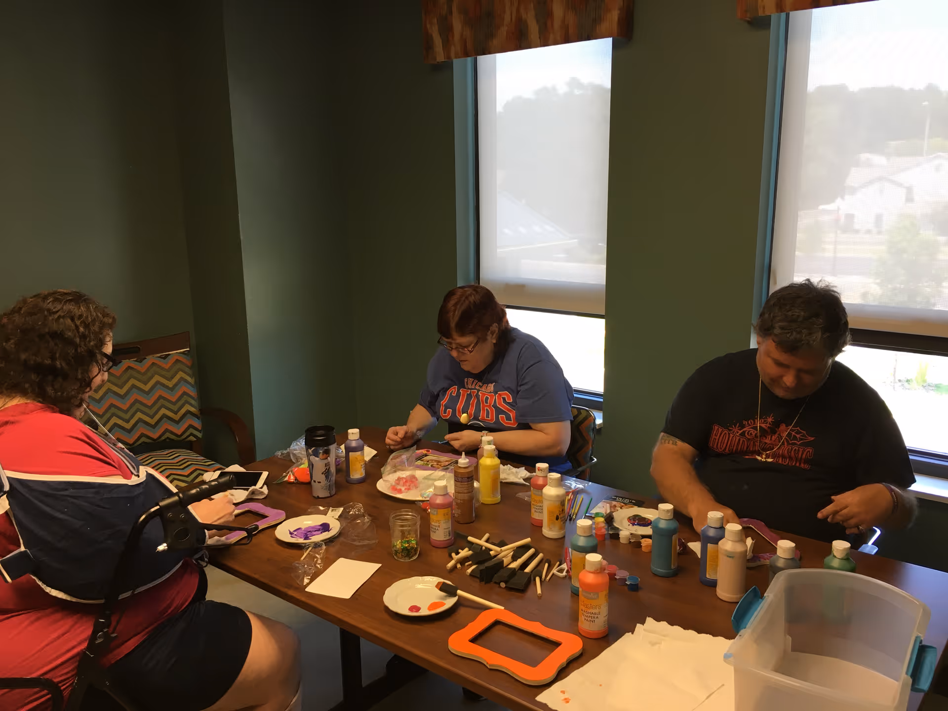 Three people sitting around a table engaged in painting crafts with various paint bottles, brushes, and craft supplies spread out on the table in a room with green walls and two windows with shades partially drawn.