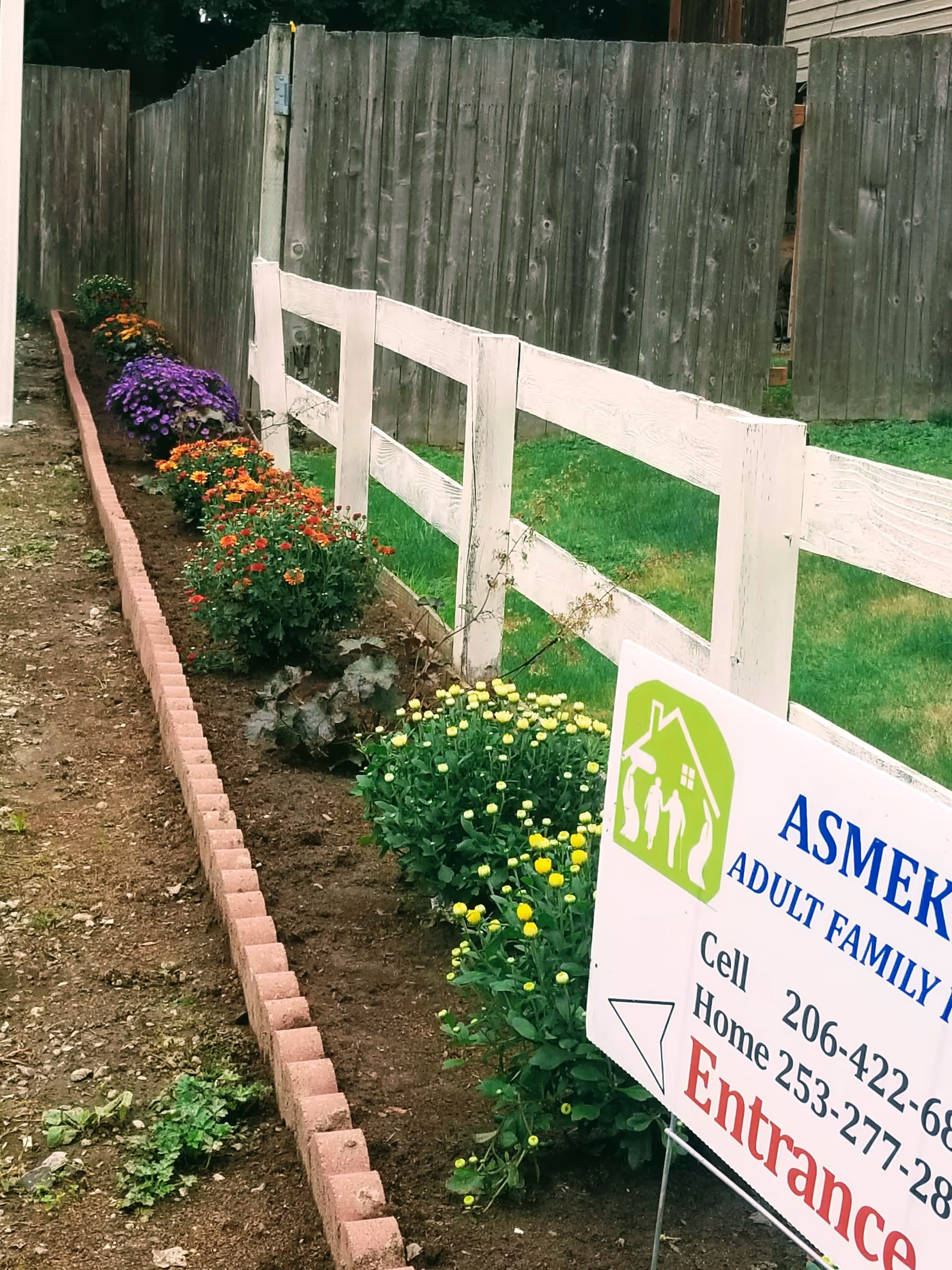 Outdoor view of a flower-lined garden beside a white fence and entrance sign.