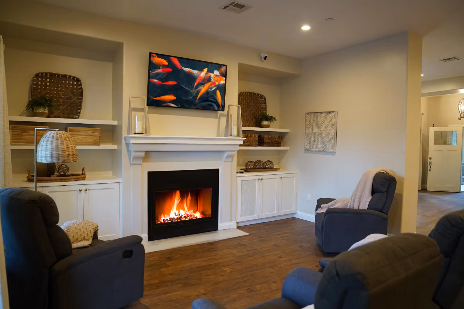 A cozy living room with a lit fireplace beneath a wall-mounted TV displaying colorful fish. The room features comfortable reclining chairs with blankets, built-in shelves with decorative baskets and plants, and wooden flooring.