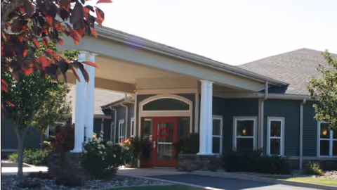 Exterior view of a senior living community building entrance with a covered porch supported by white columns, a red door with a wreath, and surrounding greenery including trees and bushes.