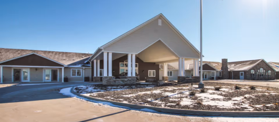 Exterior view of Cedarhurst Senior Living of Bethalto building with a covered entrance, multiple windows, and a driveway. There is some snow on the ground and the sky is clear and blue.