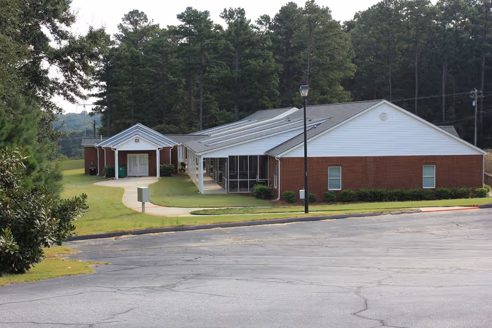 Single-story brick-and-white siding senior living building with a covered entrance, walkway, and surrounding trees.