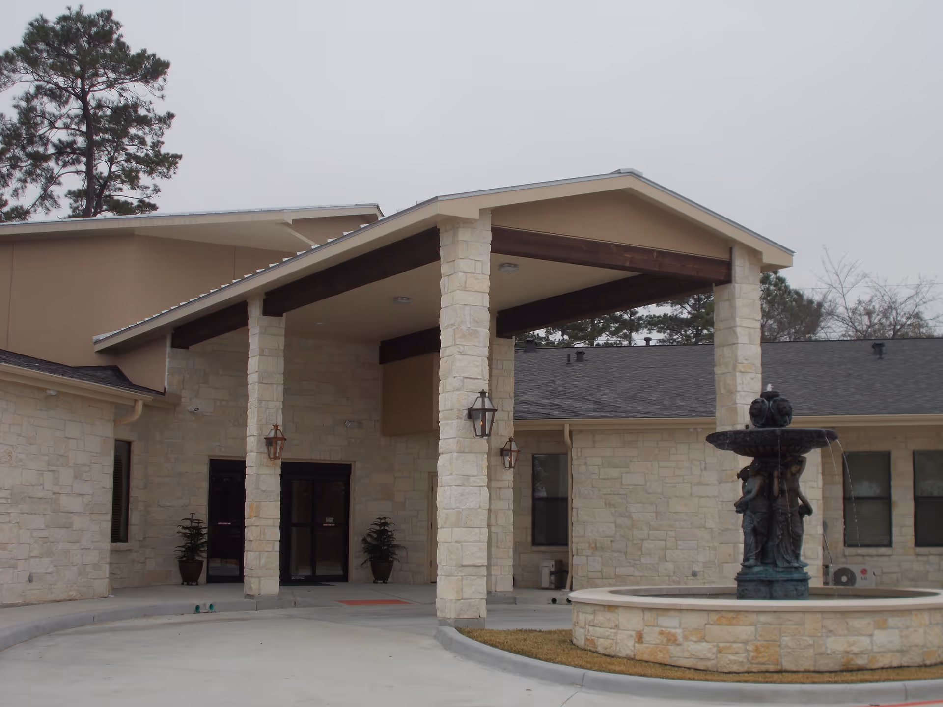 Exterior view of The Bellaire Senior Lodges building entrance with stone pillars supporting a covered driveway and a decorative fountain in front.