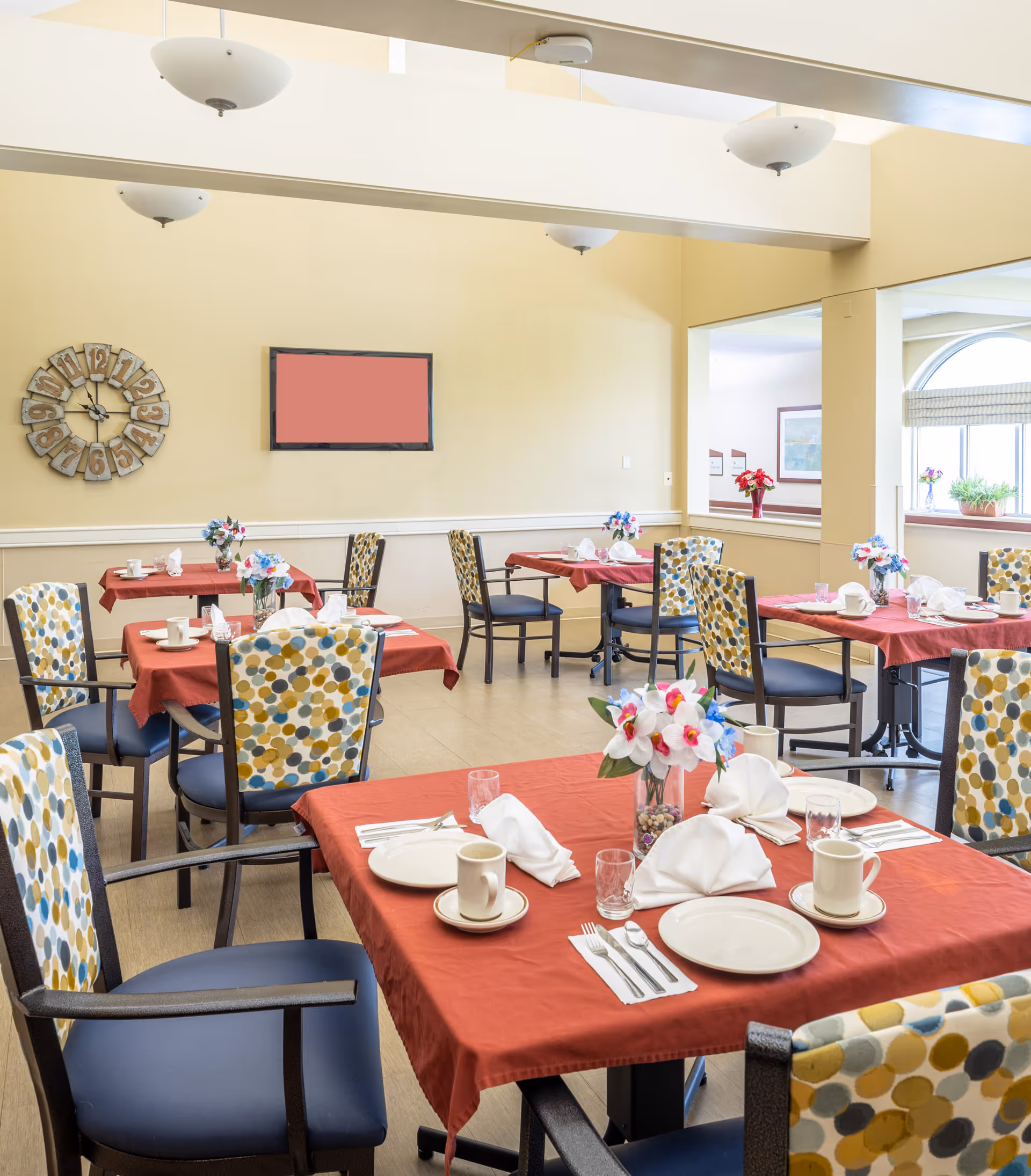 Bright dining room with tables set with red tablecloths, place settings, floral centerpieces, and patterned chairs.