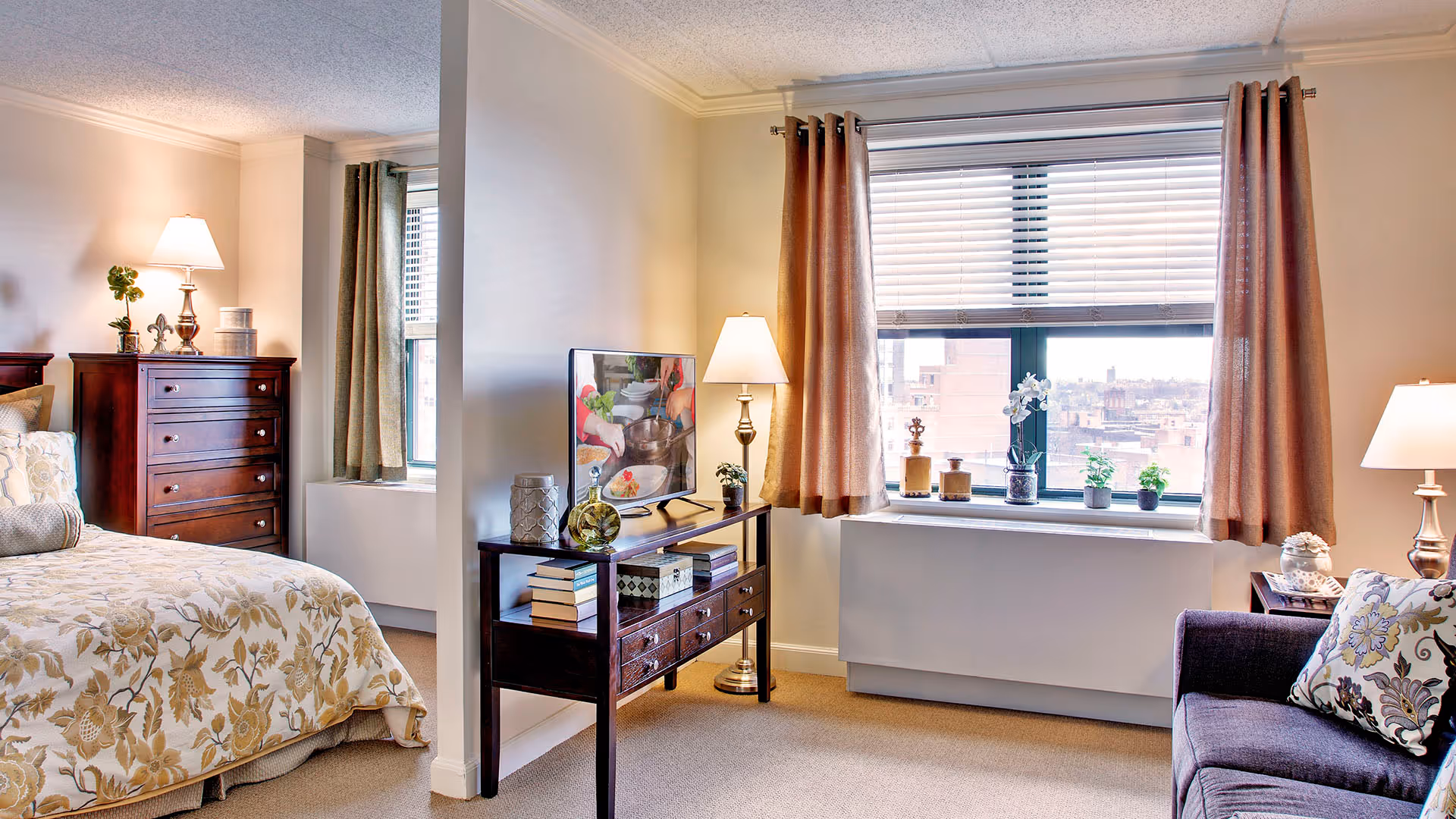 A cozy senior living bedroom with a floral bedspread, a wooden dresser, a TV on a wooden stand with books and decorative items, a window with beige curtains and potted plants on the windowsill, a purple armchair with a floral pillow, and two table lamps providing warm lighting.