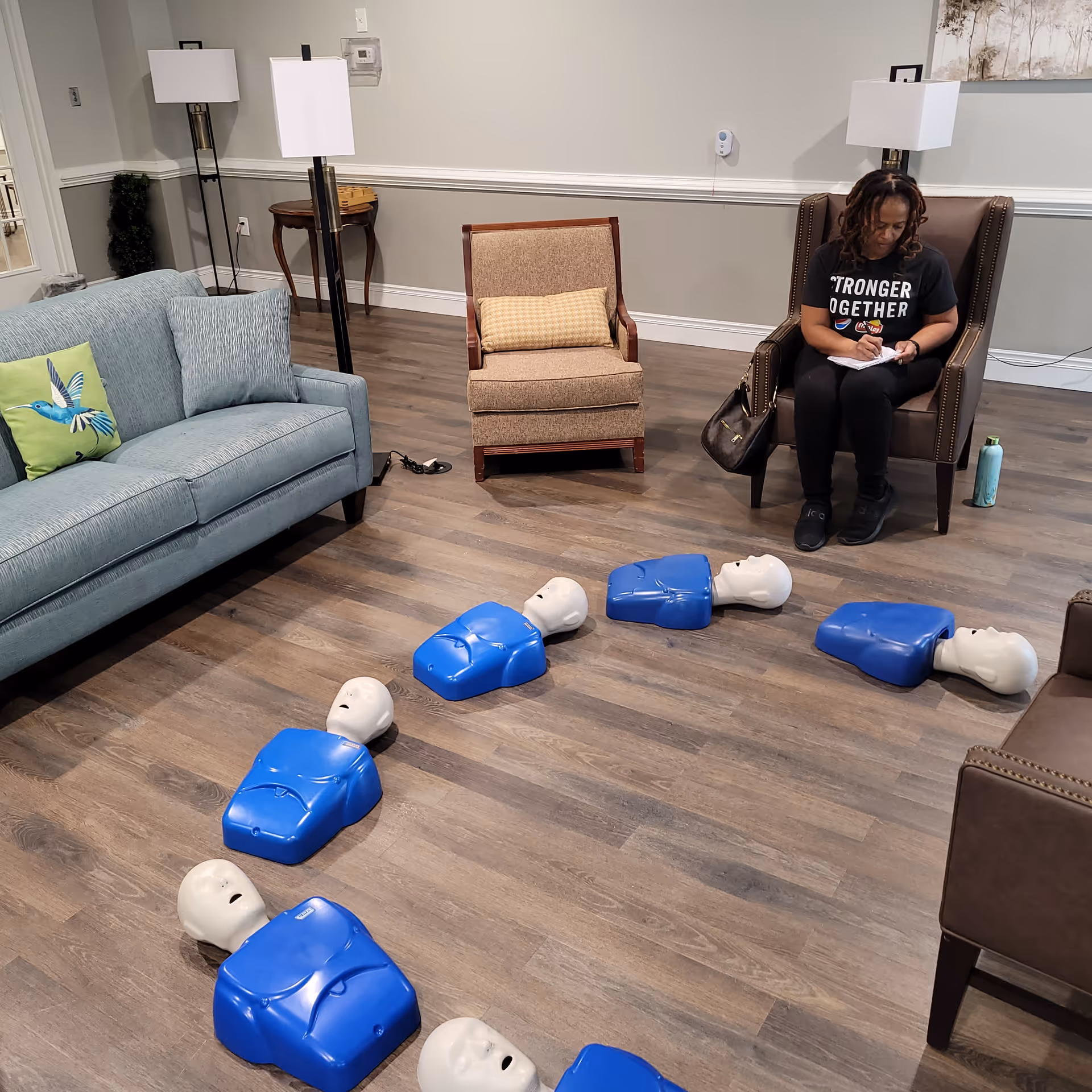 A lounge room shows CPR training mannequins arranged in a circle with a seated person.