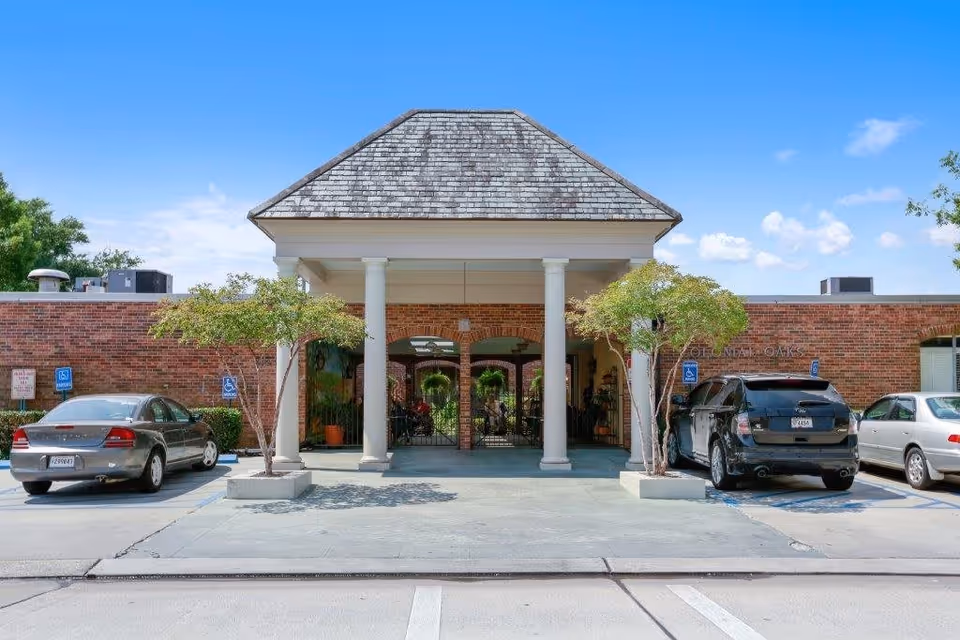 Front entrance of a brick senior living facility with a columned portico, potted trees, and parked cars in accessible spaces.