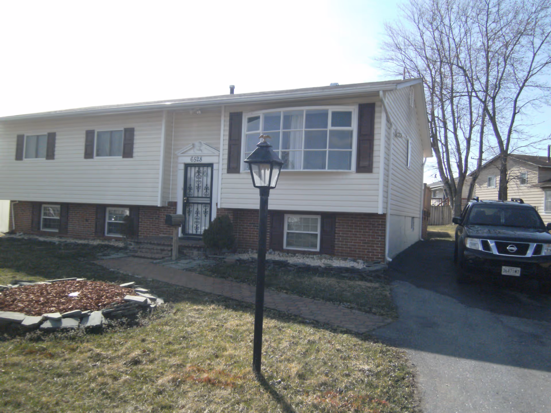 Exterior view of a single-story residential house with beige siding and brick foundation. There is a black lamp post in the front yard, a paved walkway leading to the front door, and a black SUV parked in the driveway. Leafless trees are visible in the background.