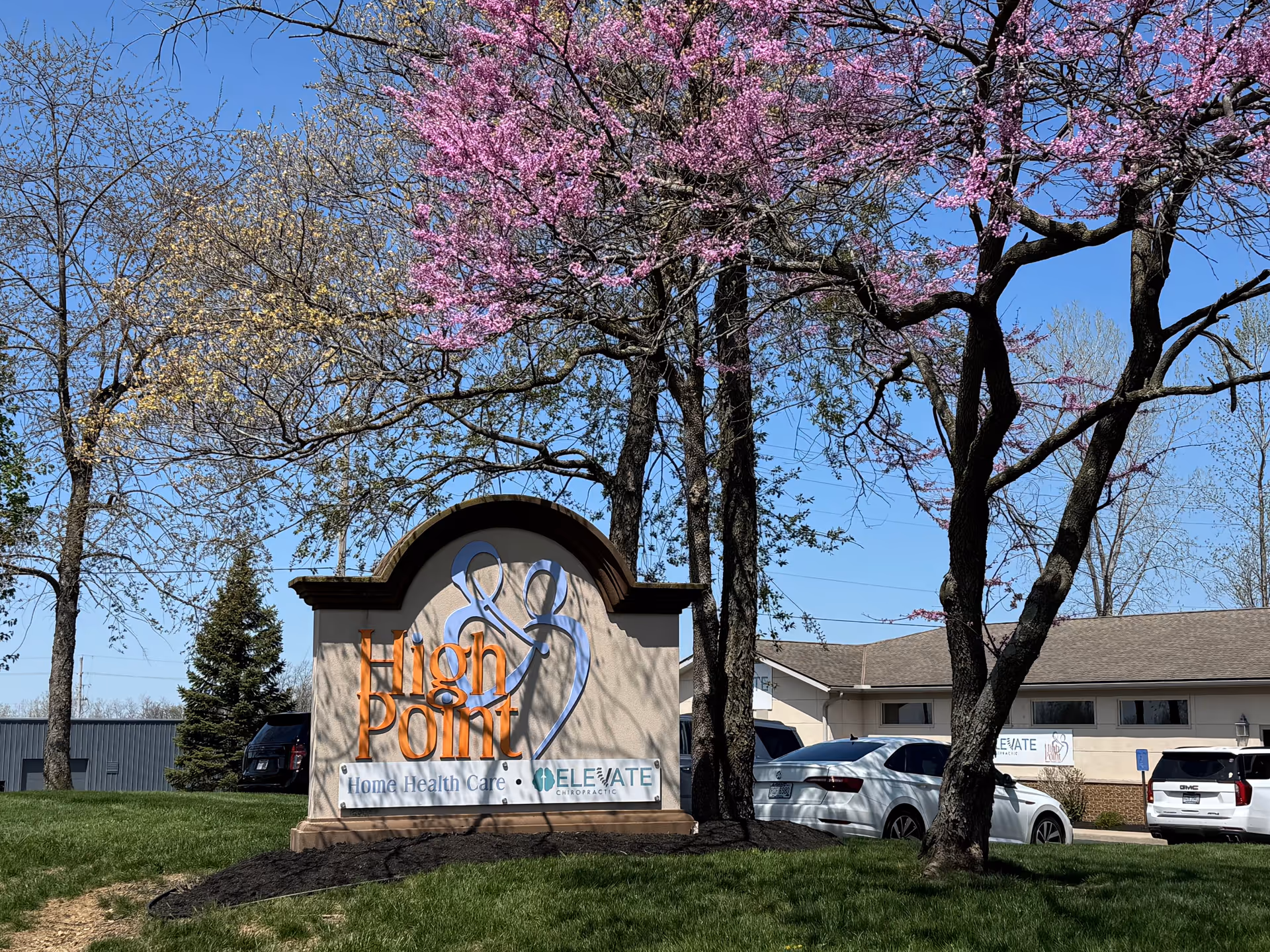 Outdoor view of High Point Home Health sign with blooming trees and parked cars.