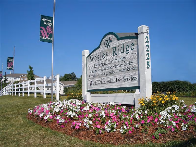 Entrance sign for Wesley Ridge retirement community next to a flowerbed and white fence under a clear blue sky.